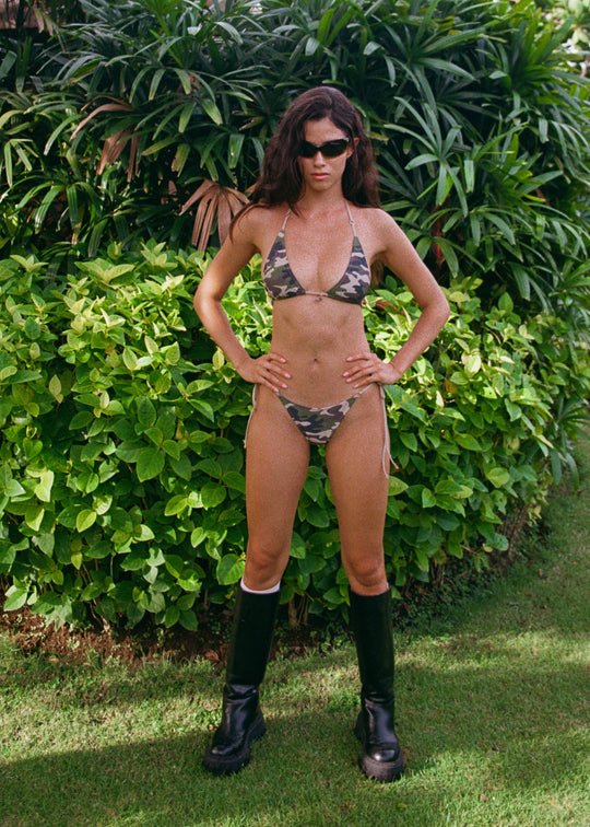 Person wearing a camouflage bikini and black boots standing in front of green foliage.